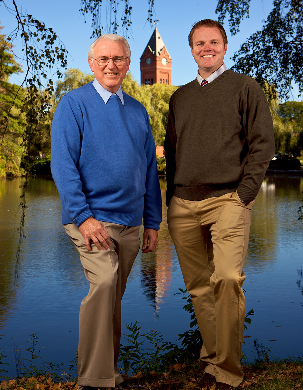 The image shows two men standing outdoors near a body of water, with one man wearing a dark blue sweater and the other in a light blue shirt. They are positioned in front of a building with a clock tower, which is part of a larger structure that appears to be a church or similar institution. The setting suggests a formal event or gathering, as indicated by their attire and the presence of a watermark or logo on the image.
