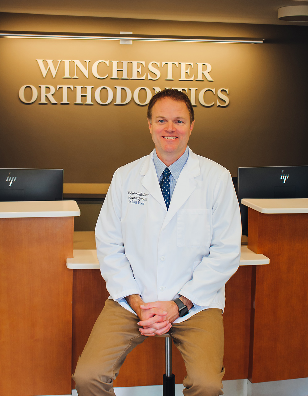 The image shows a man in a white lab coat standing at a desk with a nameplate that reads  Winston Orthodontics  behind him.