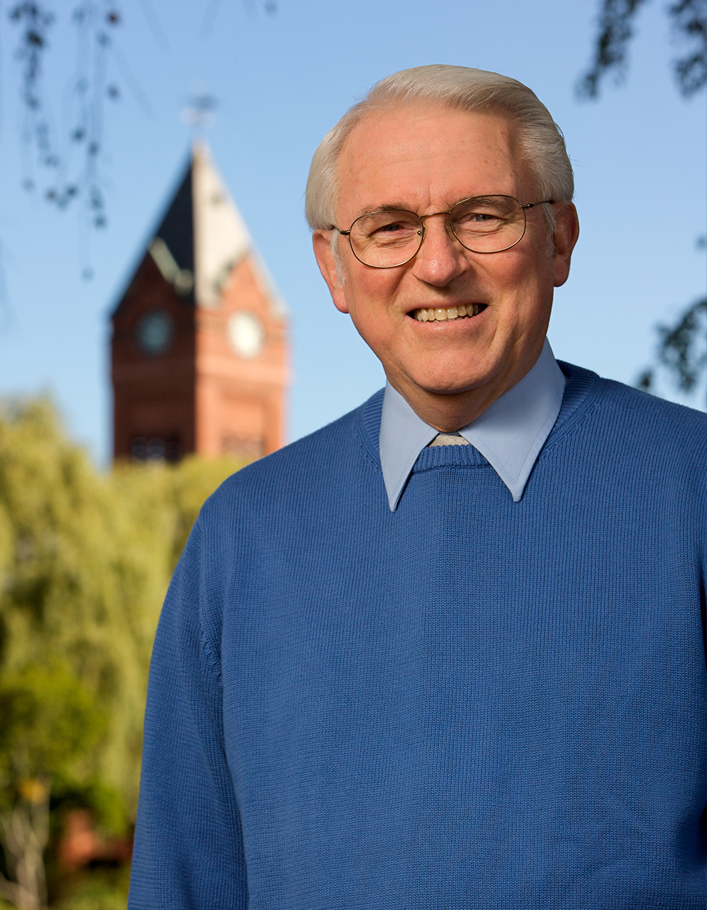 The image shows an older man standing outdoors in front of a building with a clock tower, smiling and posing for the photograph.
