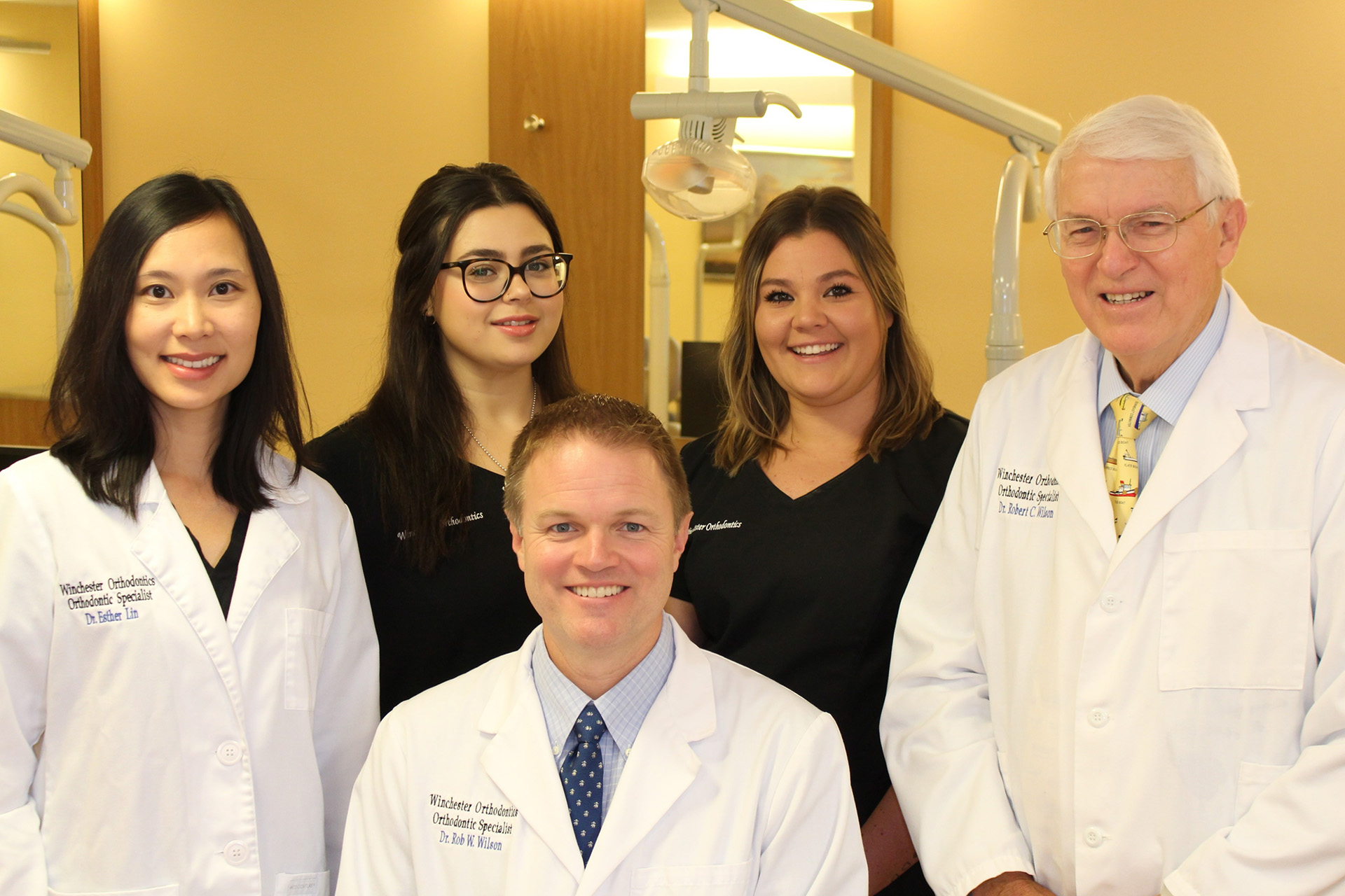 The image shows a group of five individuals, likely professionals given their attire, posing together in what appears to be an office setting. They are standing behind a man seated on the left, who is distinguished by his tie and glasses, suggesting he may hold a position of authority or expertise within the context of this photograph. The background features a mirror reflecting part of the room, indicating that the photo was taken indoors.