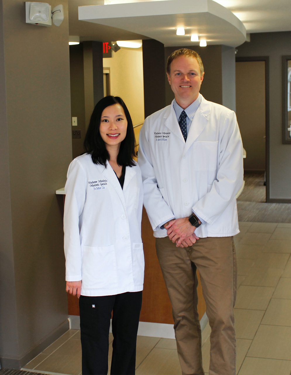 The image shows a man and woman, both dressed in white lab coats, standing together in an indoor setting that appears to be a lobby or waiting area. They are posing for the photograph with smiles on their faces.