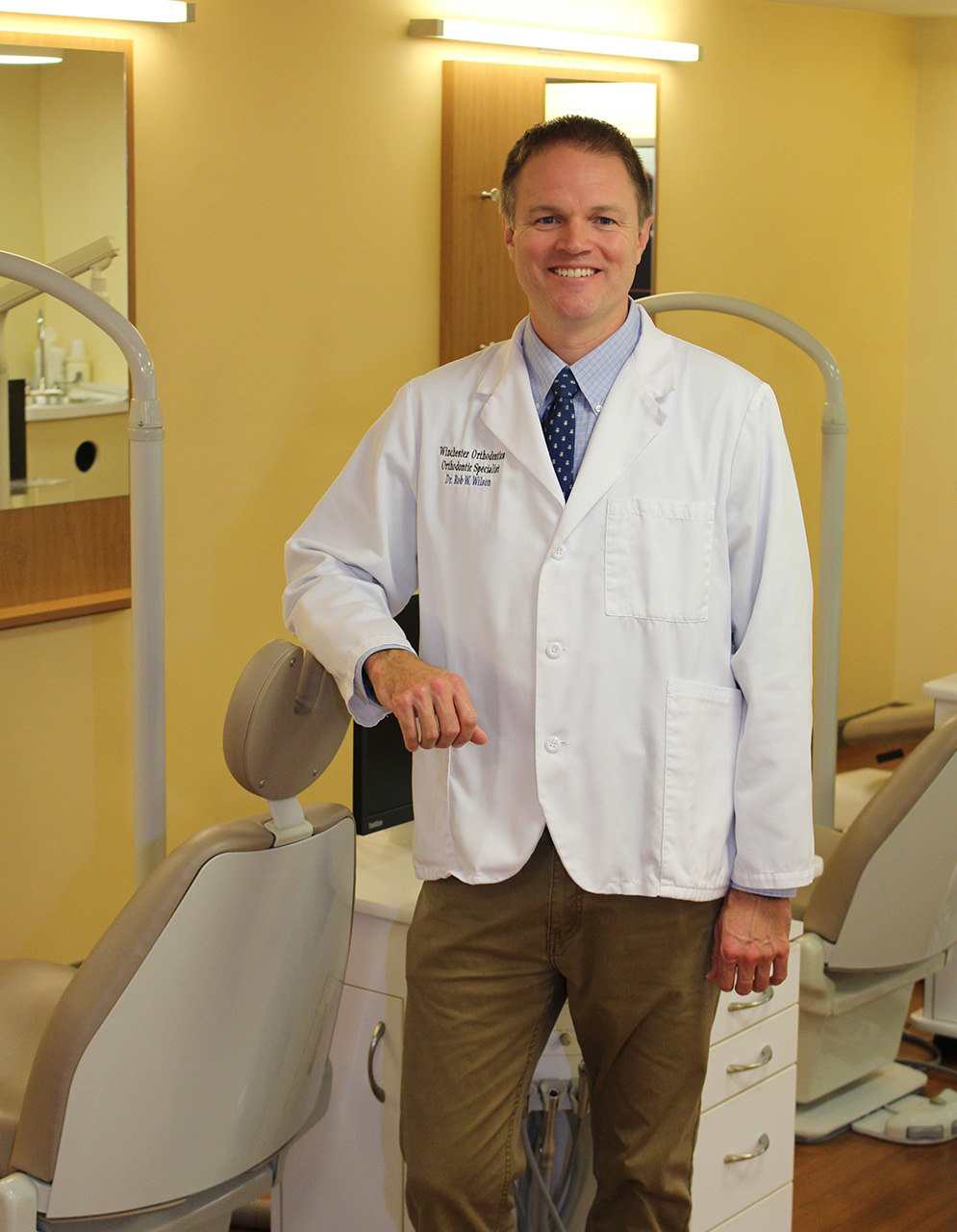 A man in a white lab coat stands confidently next to a dental chair, smiling at the camera.