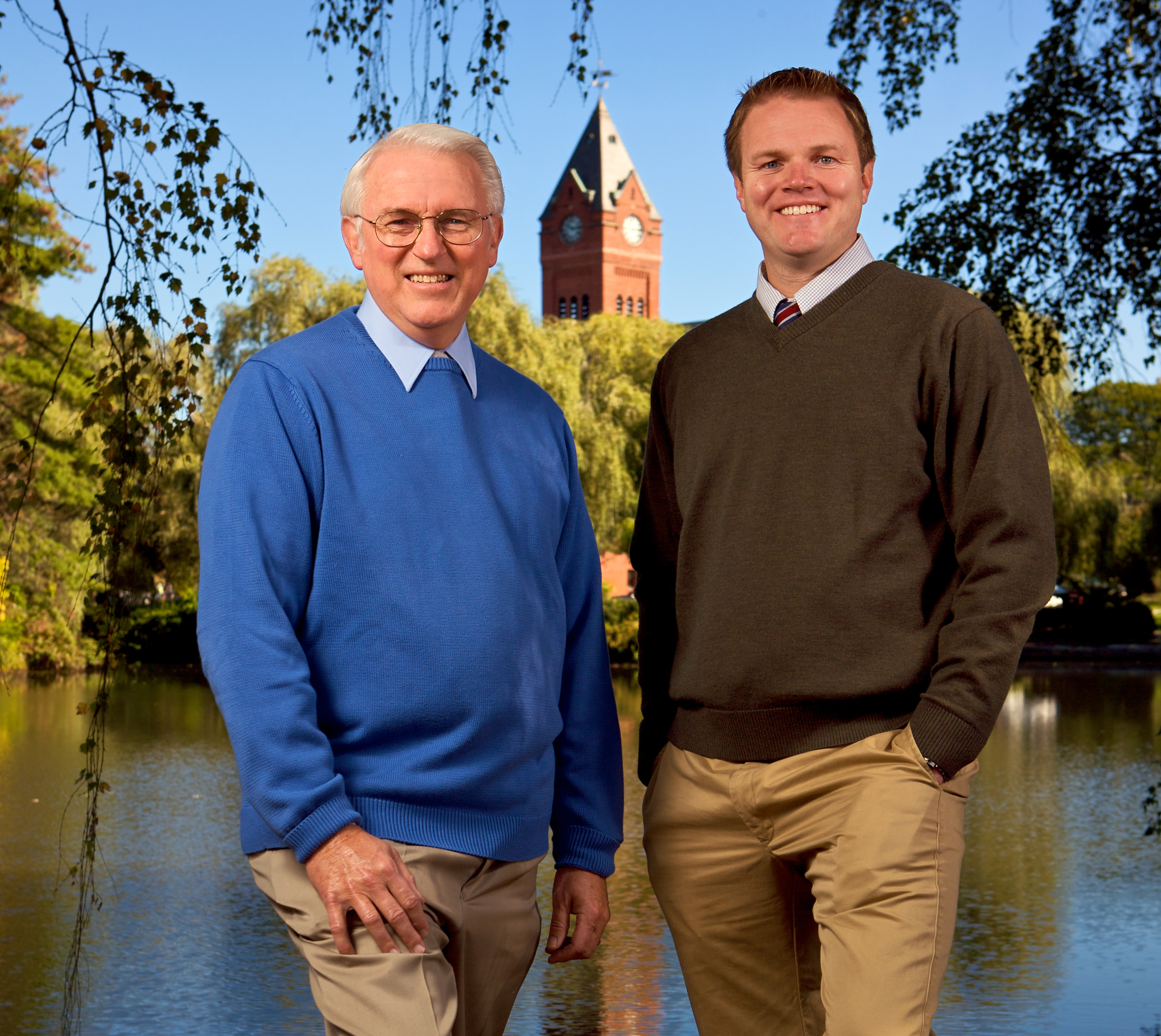 The image shows two men standing outdoors in front of a body of water, with one man wearing a blue sweater and the other dressed in a dark suit.