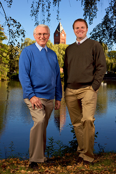 Two men standing outdoors near a body of water, with one man dressed in formal attire and the other in casual clothing.