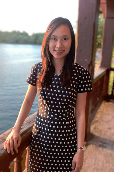 The image shows a woman standing outdoors at sunset, posing for the camera with a smile. She is wearing a polka dot dress and stands on a wooden deck overlooking water.