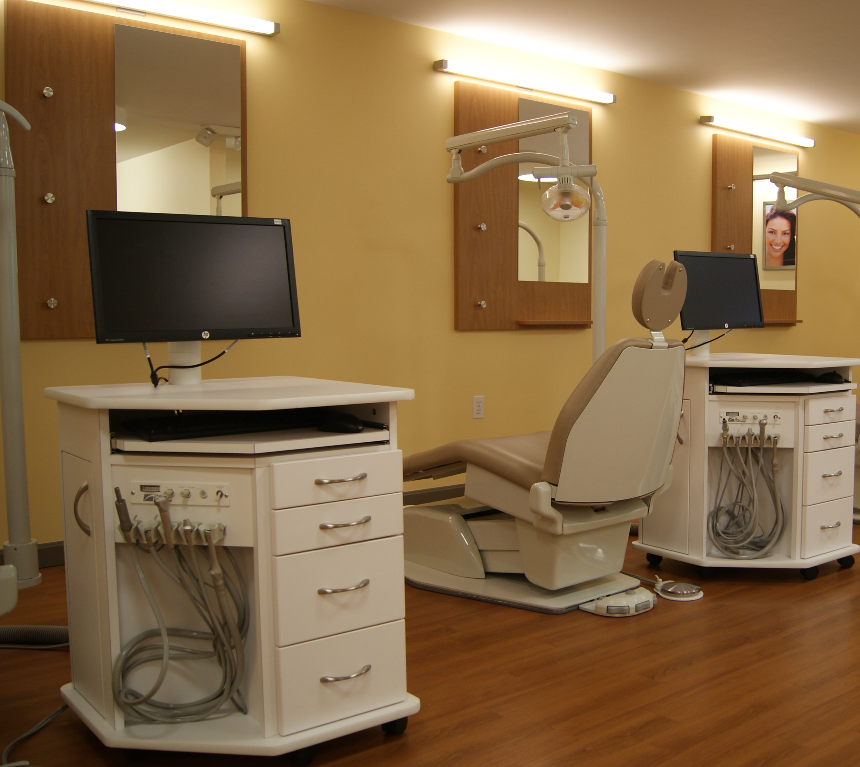 The image shows a dental office interior with modern equipment, including a chair for dental examinations and a computer monitor.