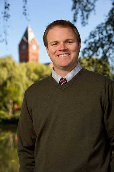 The image shows a man standing outdoors, dressed in a dark sweater and tie, smiling at the camera. Behind him is a clear sky, green trees, and a building that appears to be a church with a clock tower.