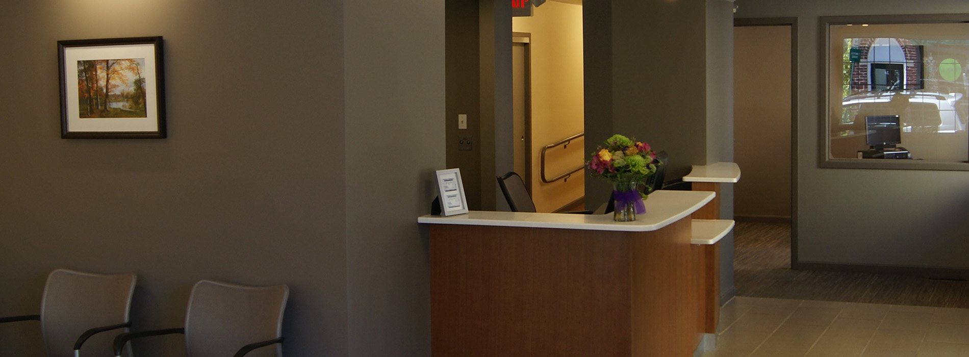The image shows an interior view of a waiting area in a building, featuring a reception desk with a sign and a seating area with chairs.