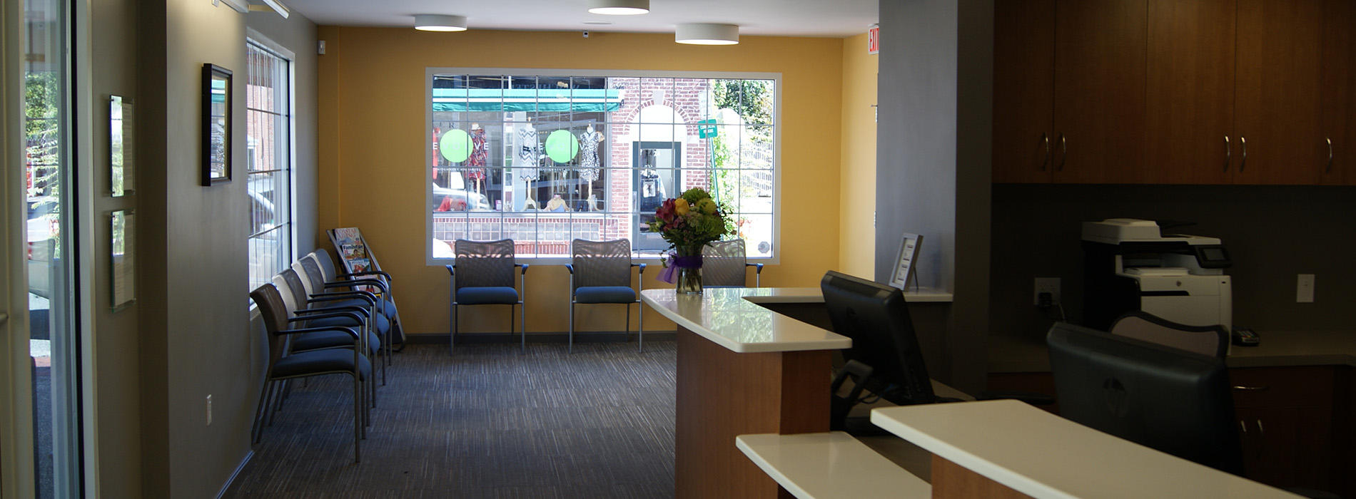 The image is a composite of two photographs showing an interior space, likely a waiting area or reception room in an office setting. The top photo shows a view from the hallway into the room with a large window allowing natural light to enter and a green color scheme on the walls. The bottom photo presents the same room but taken from a different angle, showcasing a desk and chairs against one wall.