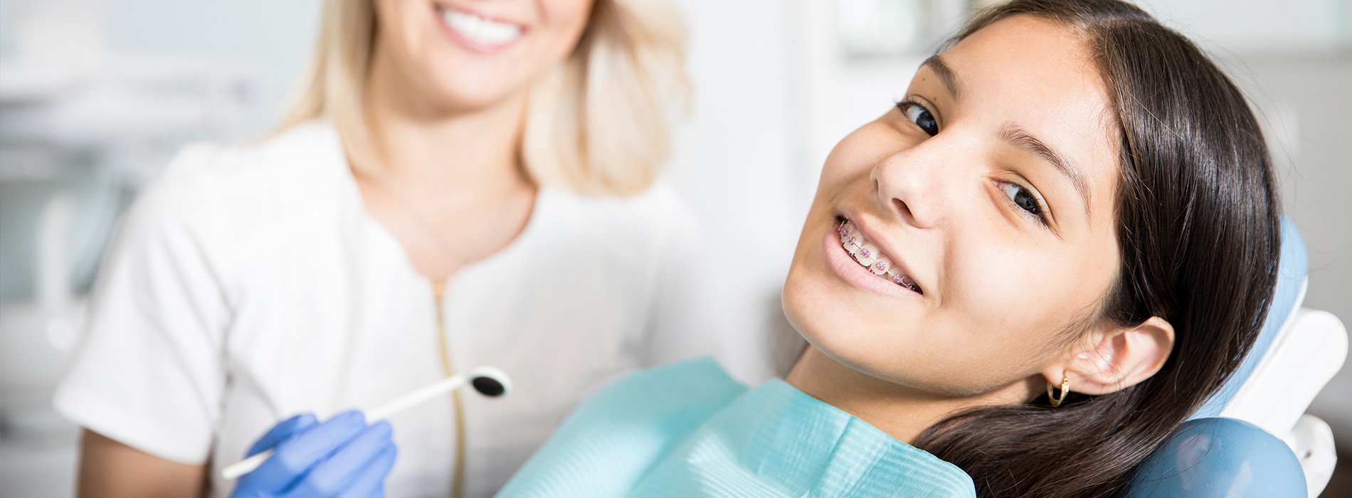 Woman in white shirt smiling at camera, sitting in dental chair with dental hygienist standing behind her.