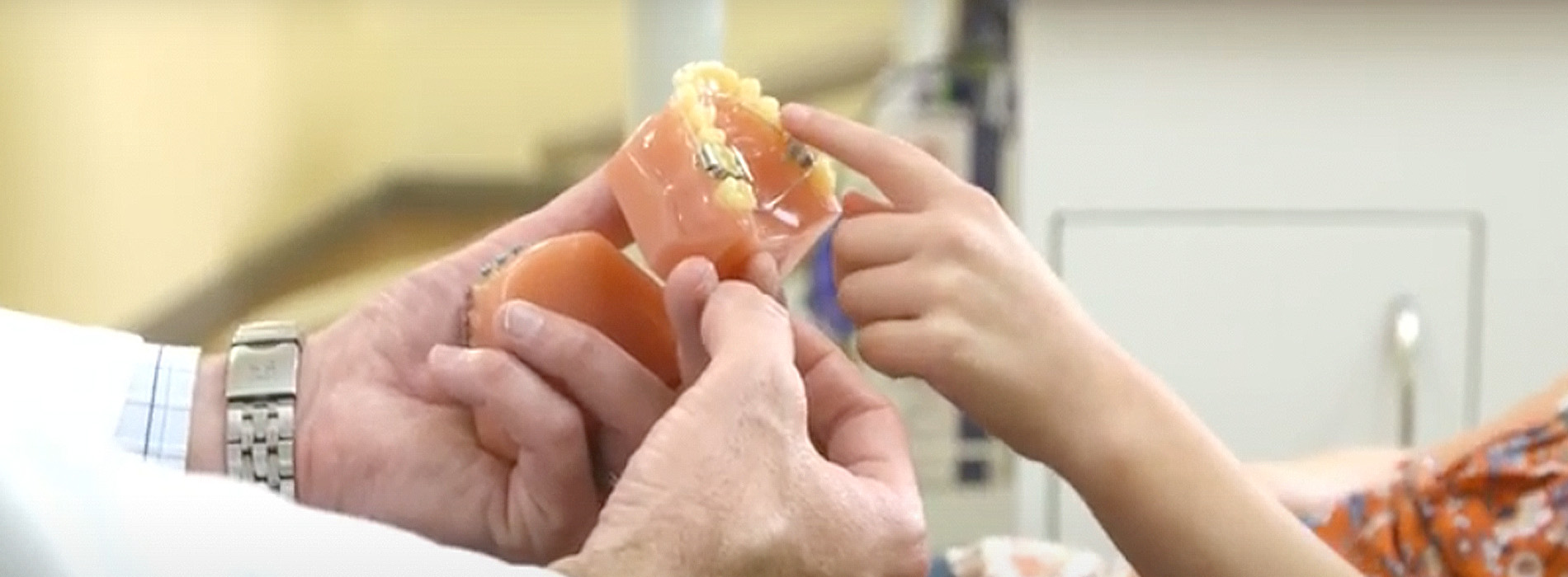 The image shows two people, one adult and one child, holding a small orange object between them. They are in an indoor setting with a blurred background that suggests it might be a kitchen or a room with cooking equipment.