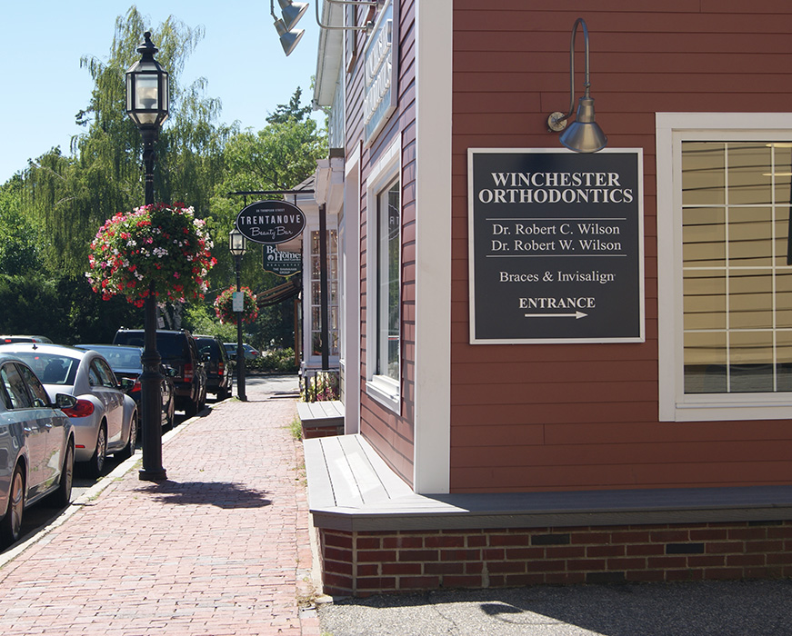 A storefront with a sign that reads  Winston Orthodontics  and a sign for an entrance, located on a sidewalk next to a parking area in a town setting.