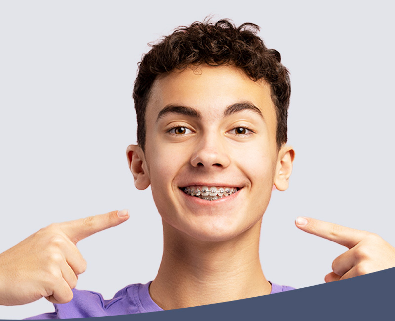 The image features a young individual with short hair, smiling broadly at the camera. They are pointing to their teeth with one hand while holding up an index finger with the other, suggesting a playful or confident demeanor.