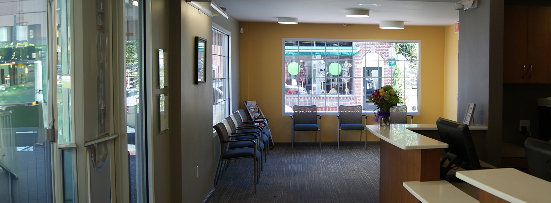 The image shows an interior view of a modern lobby or waiting area with a neutral color palette, featuring a reception desk, seating areas, and a large window.