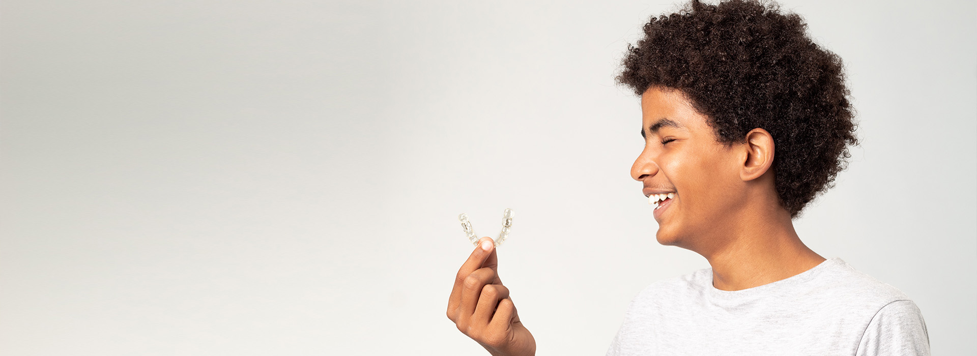 The image is a photograph of a young person holding a flower, smiling and looking to the side with one hand raised near their face. They appear to be outdoors in daylight.